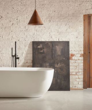 A bathroom with white exposed bricks, a copper cone light, a curved white bath tub, and a rectangular metal sheet