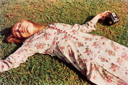 View of Marcia Hare wearing a pink floral dress lying on grass holding a black Kodak camera during the day