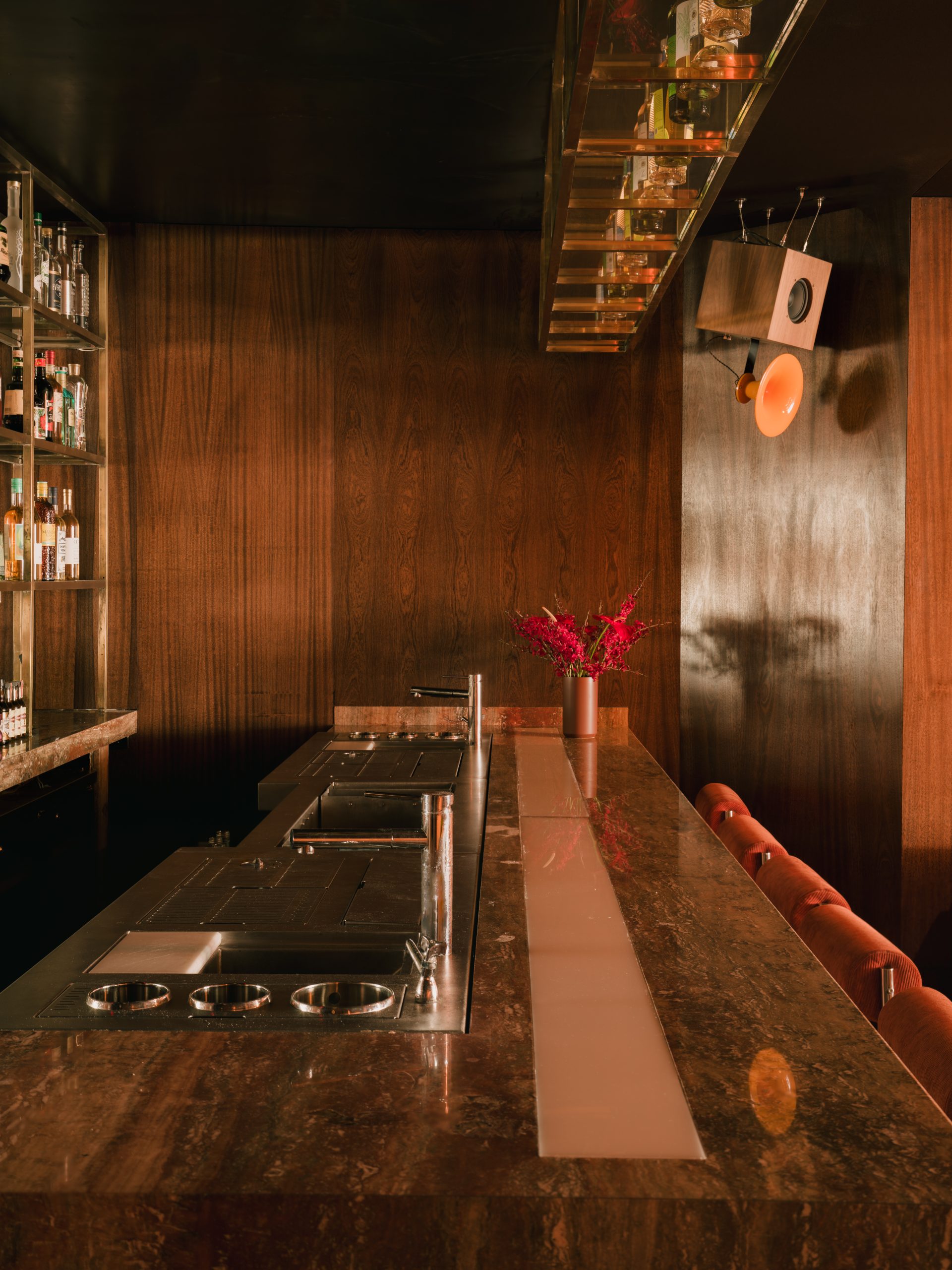 A dimly lit bar with red and orange-tinted interiors sculpted in wood, chrome, lacquer, velvet, and marble features sinuous banquettes, atmospheric sconces, geometrical skylights, and refined details.