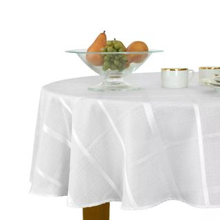 A circular table with a white tablecloth with a jacquard print, brown legs, and a glass bowl of pears and grapes in it and two white mugs with gold trims next to this