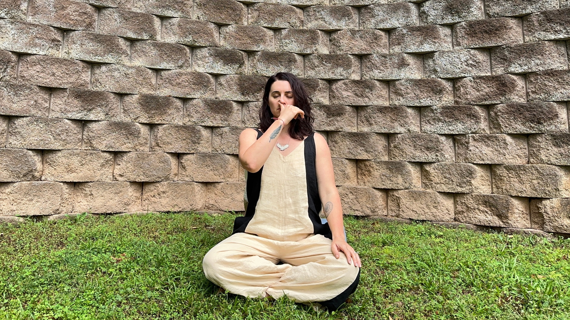 woman sat cross legged on some grass with a large stone wall behind her. she has her right thumb over her right nostril.