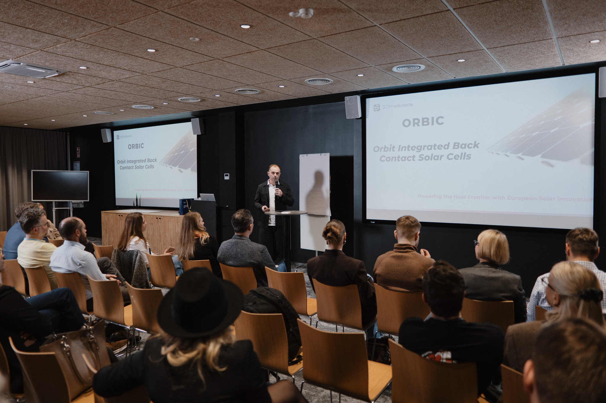 People in the audience listen to a man speaking. There is a powerpoint presentation that says "ORBIC."