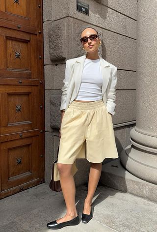 A woman wearing a white blazer over a white t-shirt with butter yellow shorts and black ballet flats.