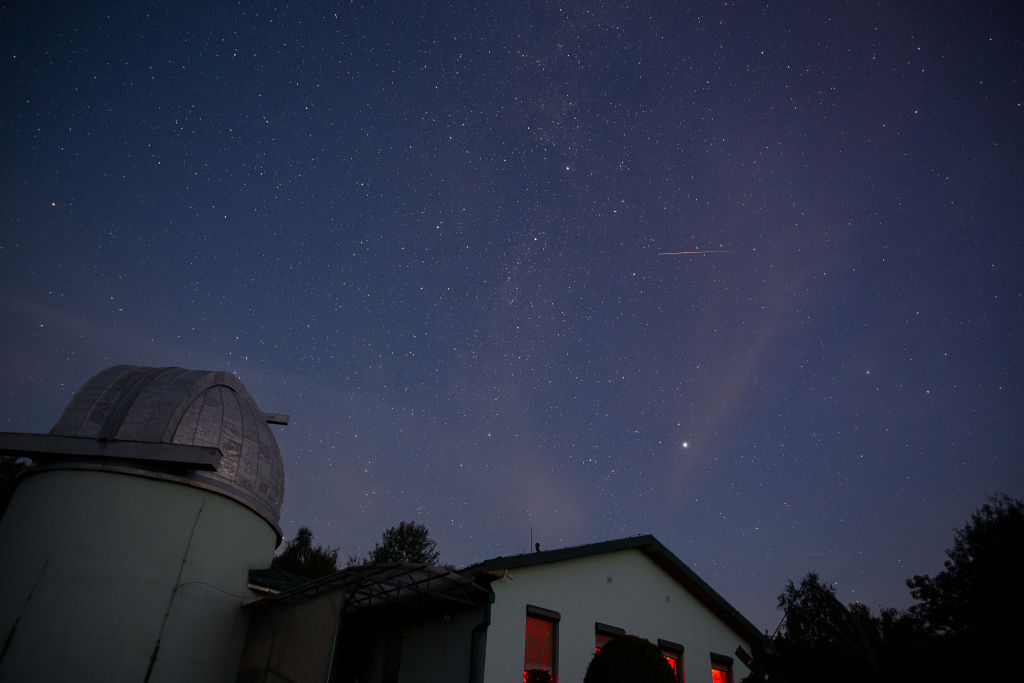 En primer plano hay un edificio blanco y a la izquierda un pequeño observatorio. El cielo está lleno de estrellas y una larga línea blanca del meteoro Perseida aparece en el cielo.