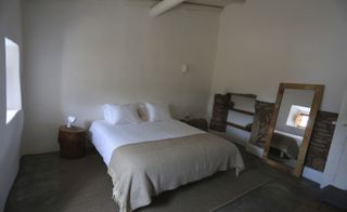 Bedroom with bed with white linen on brown rug. The walls are painted white and there is a wooden mirror on the floor against the one wall.