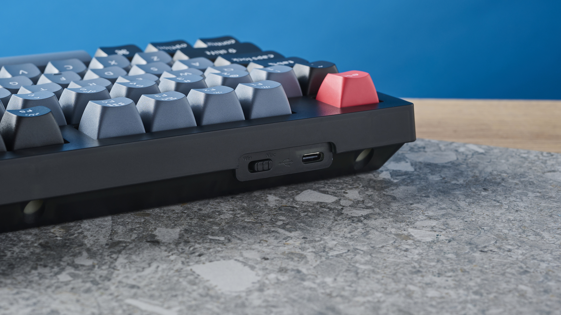 A photograph of the Keychron V6 in black, with gray , black and orange keycaps. The keyboard is positioned on a stone slate, with a blue wall in the background.