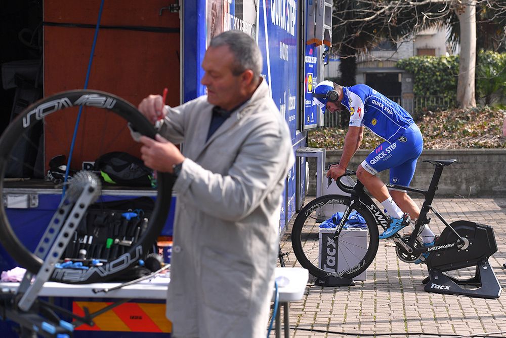 A Quick-Step mechanic glues a tyre while Fernando Gaviria rides a trainer in the background.