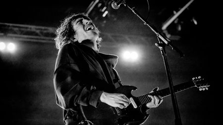 A low-angle, black-and-white photo of Cameron Winter passionately singing and playing an electric guitar onstage under bright stage lights. 