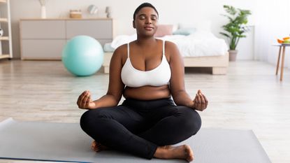 Woman in white tank top and black leggings sat meditating on yoga mat in living room