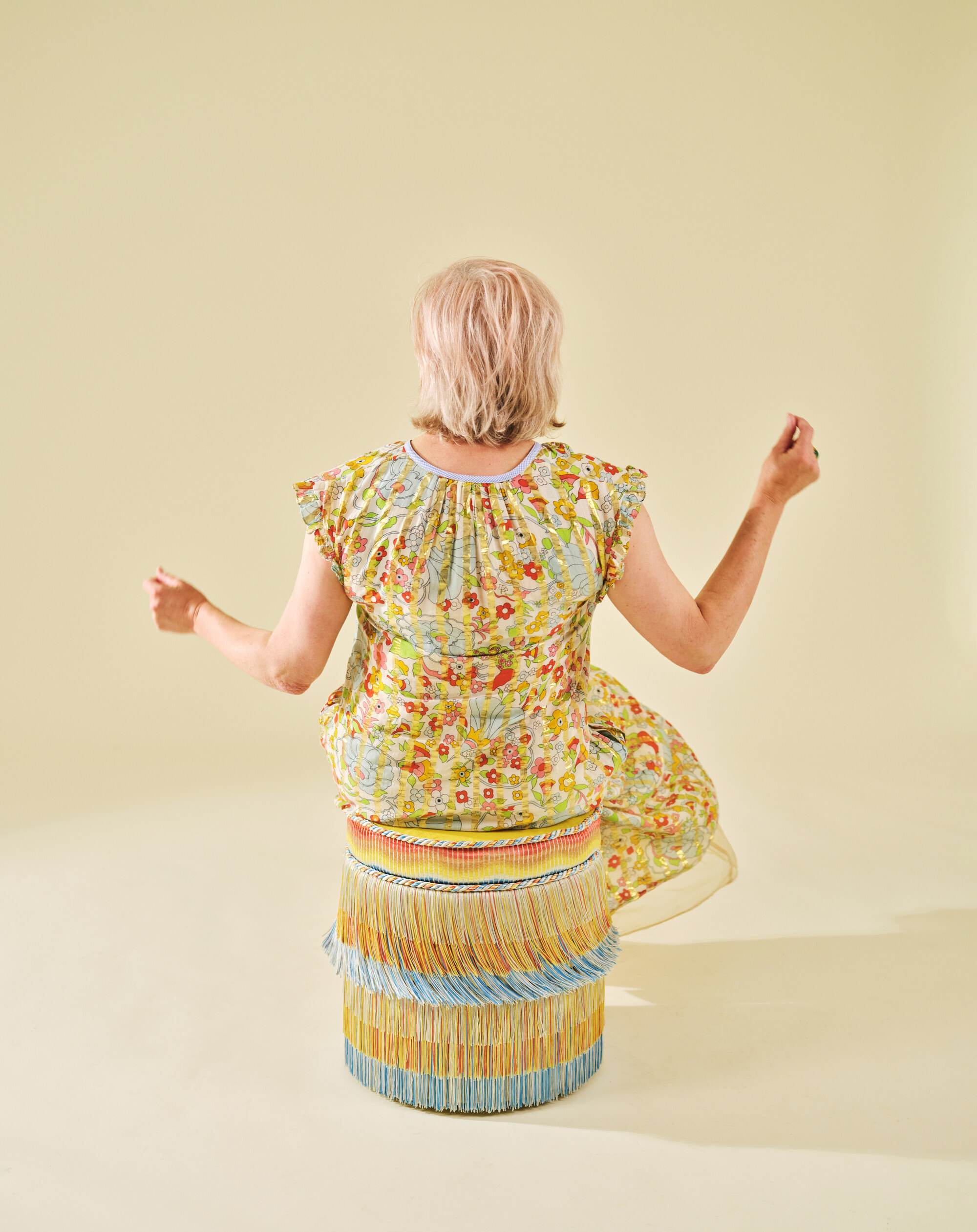 image of a woman in a yellow floral dress sitting on a stool that is blue and yellow with fringe. The background is all butter yellow