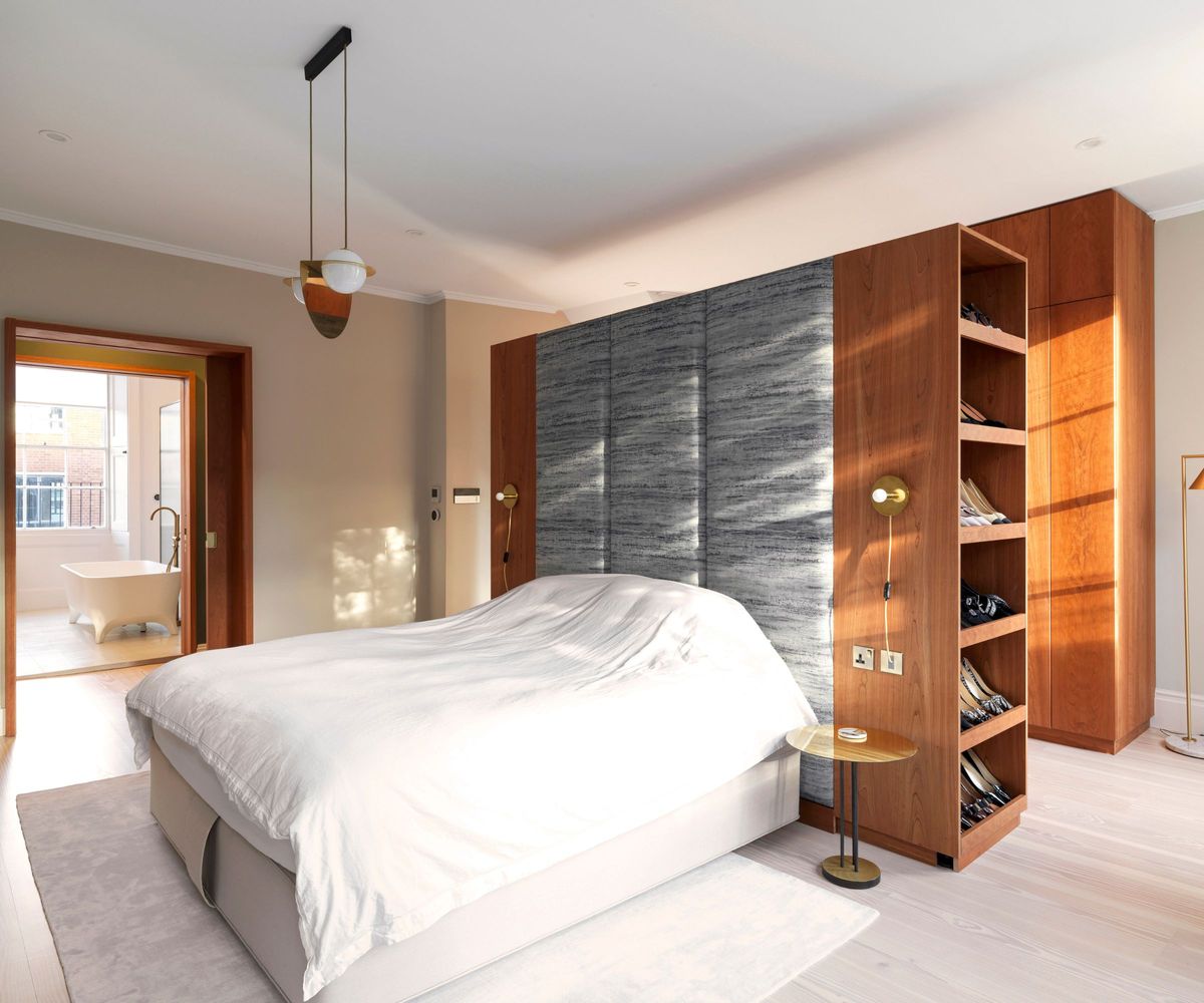 A bright bedroom with a large bed dressed in white linens, backed by a tall grey upholstered headboard. Built-in wooden wardrobes with open shoe shelves frame the space. An open doorway reveals a freestanding bathtub in a light-filled bathroom.