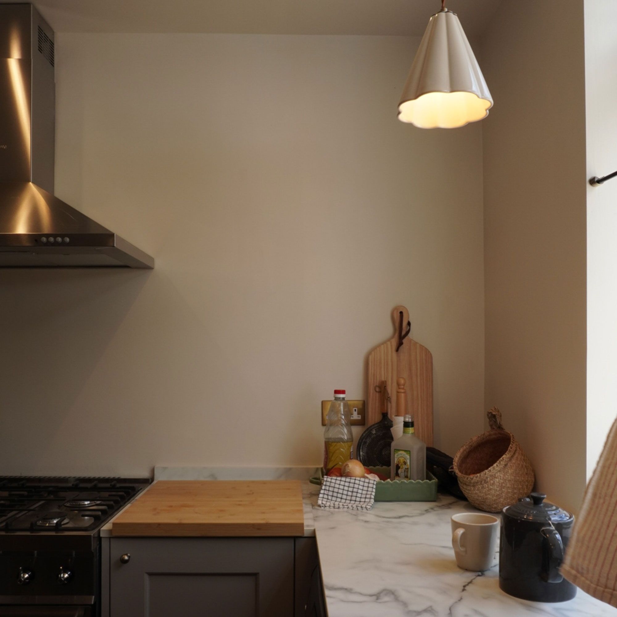 Corner of kitchen with grey cabinetry, neutral walls and marble style worktops beneath scalloped pendant light