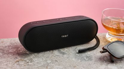 Tribit XSound Plus 2 on gray surface with sunglasses and half-filled glass with ice to its right