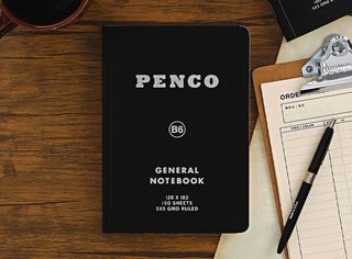 A black, portable notebook's cover reads "PENCO B6 GENERAL NOTEBOOK" all in white, while sitting on a wooden desk opposite an order sheet and other utensils.