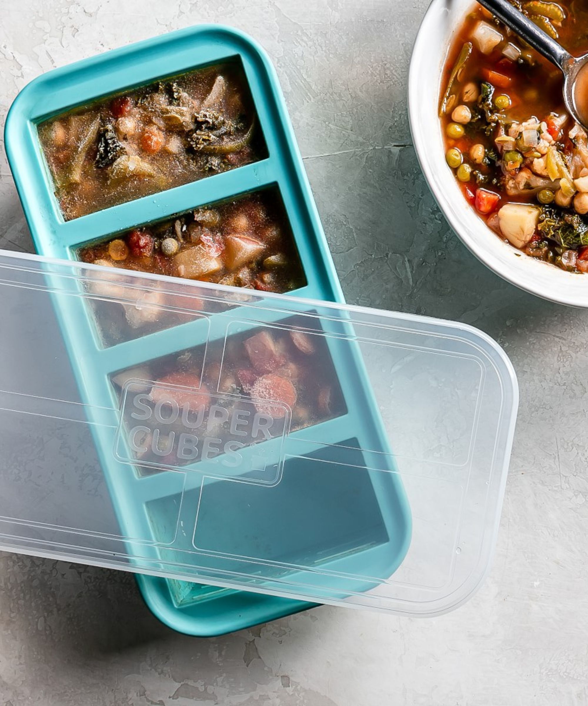 Blue Souper Cube (four sections) with frozen stew and a bowl of defrosted stew. The transparent BPA-free plastic lid is set askew on top of the Souper Cubes