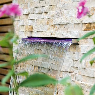 Garden water feature coming out of a brick effect wall with flower stalks in front of it