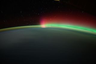 a wash of green aurora, crowned in red, wisps over the top of a night-shadowed earth.