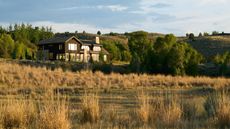 A modern stone and wood house surrounded by tall grasses and trees, set against rolling hills at sunset