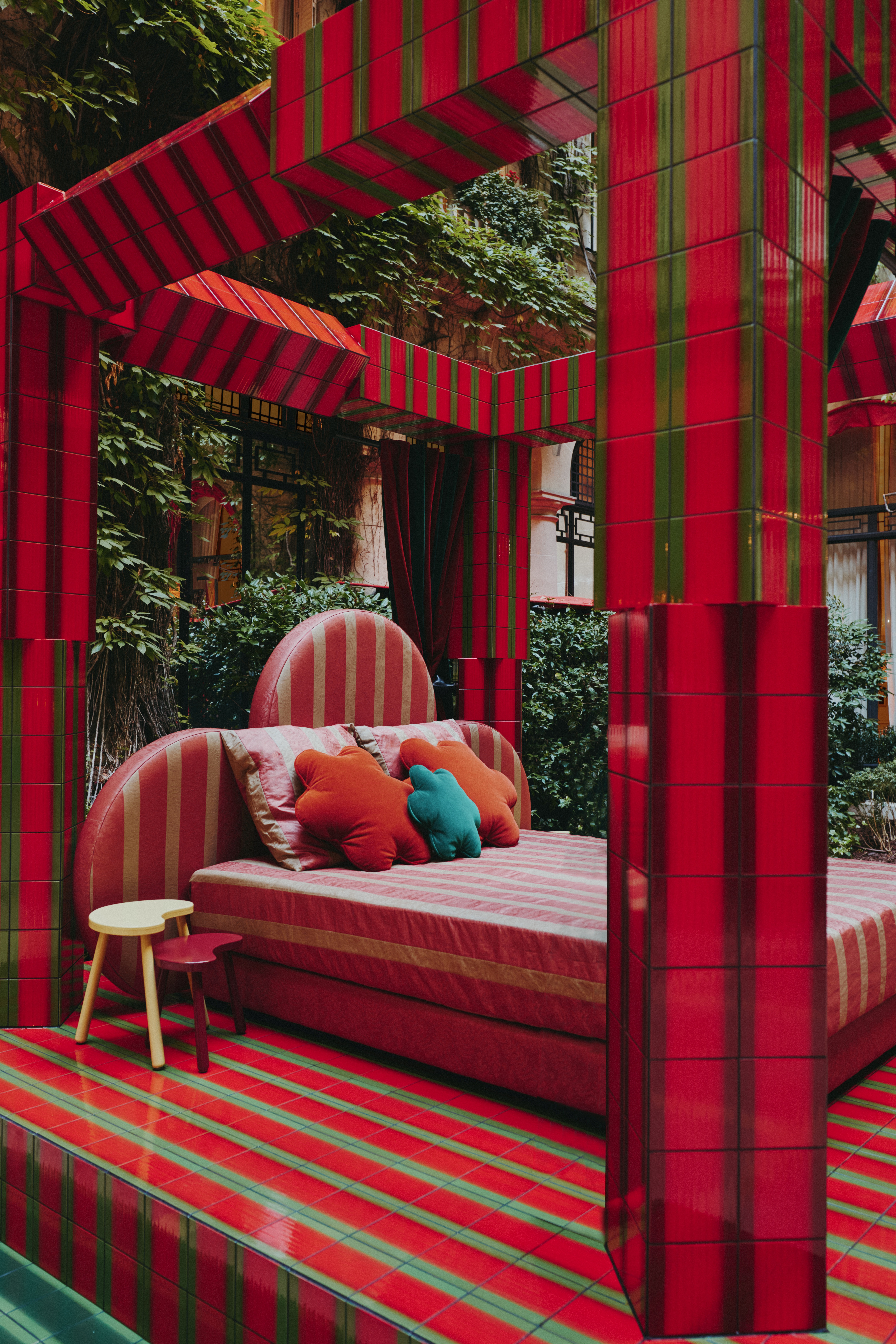 A red and green, tiled bed canopy sits in a leafy inner courtyard while platforming a bed with a clover-shaped headboard, floral cushions in red and green, and gold and red stripy sheets.
