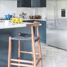 Kitchen with blue kitchen cabints and a blue kitchen island, with a bar stool in front of it. There's also grey herringbone flooring and a big fridge/freezer