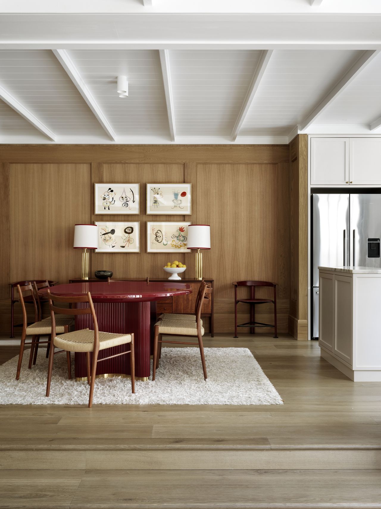 Image of a wood-paneled dining room that opens into a white kitchen. In the dining area, there is a lacquered red dining table with wooden chairs around it and a fluffy white rug on the ground.