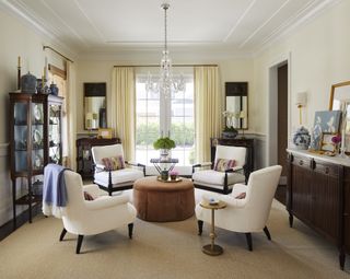 traditional living room with chandelier, four white armchairs styled around a round coffee table, a console table and cabinet against either wall, and a window looking out to the garden