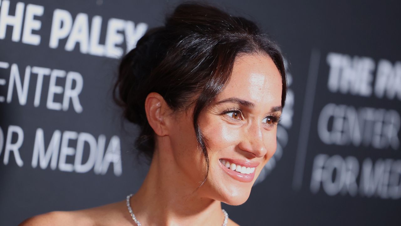 A headshot of Meghan Markle wearing a diamond necklace and a curly updo in front of a black Paley Center for Media background