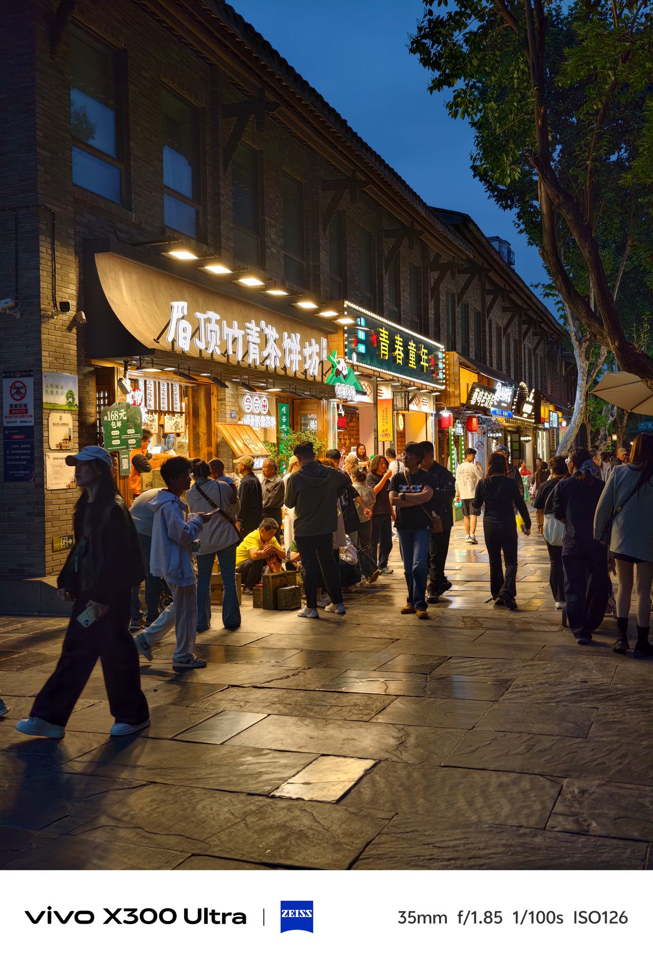 Pedestrians walking past brightly lit shops with Chinese signage on a stone street at dusk.
