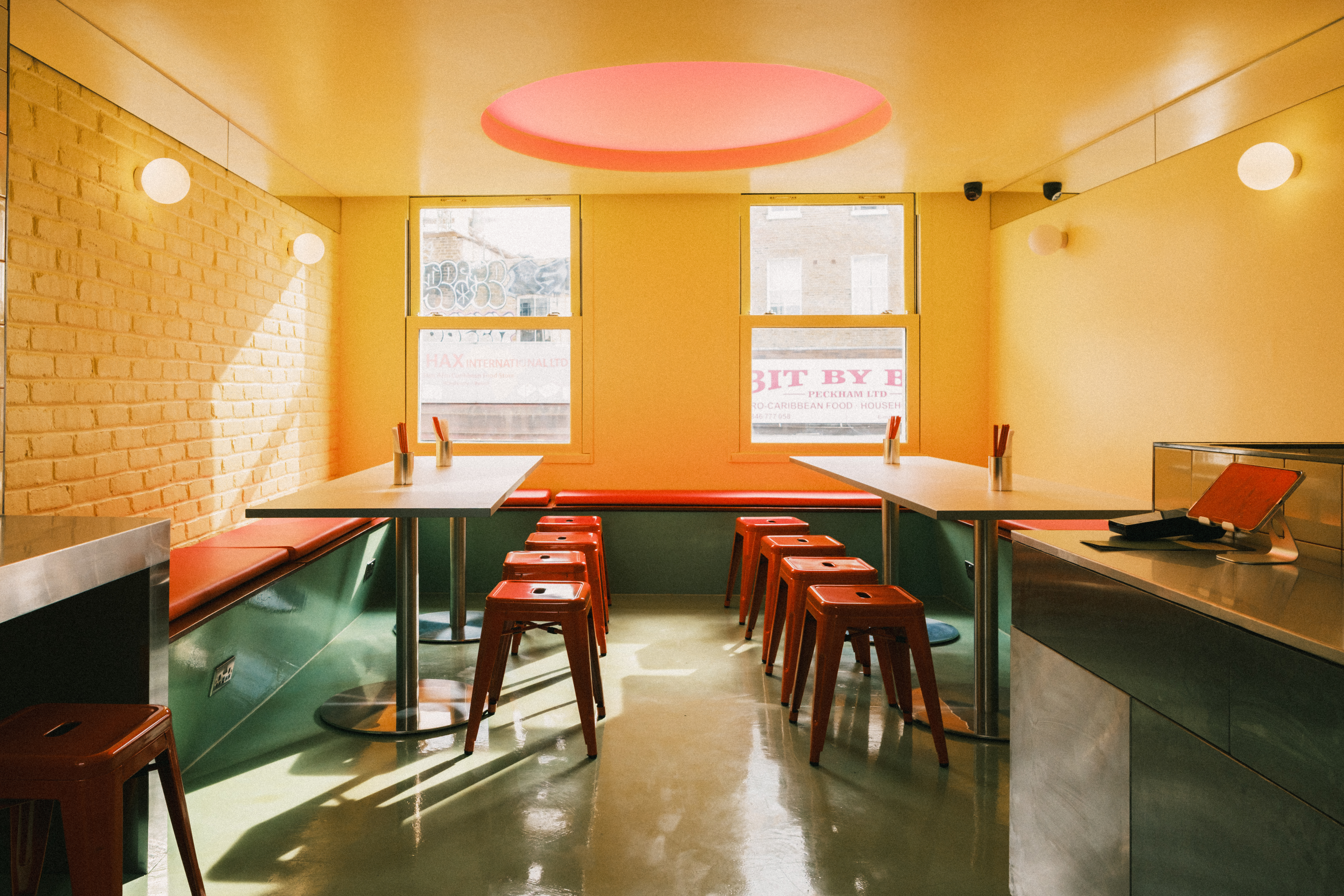 A lively, small restaurant features buttery yellow walls made of bricks, mint green floors, silver counters, and wood communal tables paired with lacquered red stools, two spherical sconces in glass and a central porthole light in red illuminating the space.