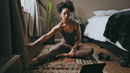 A woman stretching legs at home, bedroom or house on floor with bed curtains in front of laptop for online fitness tutorial.