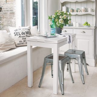 small dining space with table with leaves, pair of galvanised stools, window seat, cushions, white dresser, neutral tiled floor