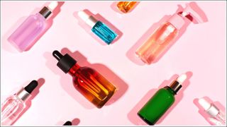 Multi-coloured glass skincare bottles and serums with shadows on a pink background.