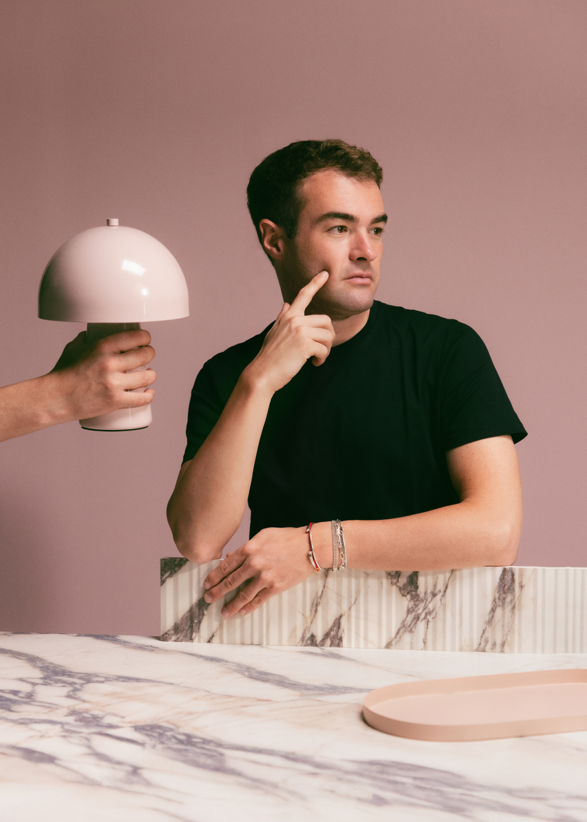 A man in a black t-shirt standing against a panel and a marble table with a pink lamp held up near him and a pink tray on the surface