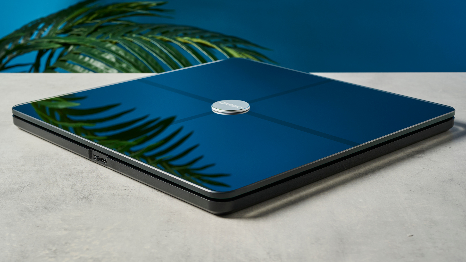 a modern black smart scale with a silver circle in its center rests upon a light surface with a blue background