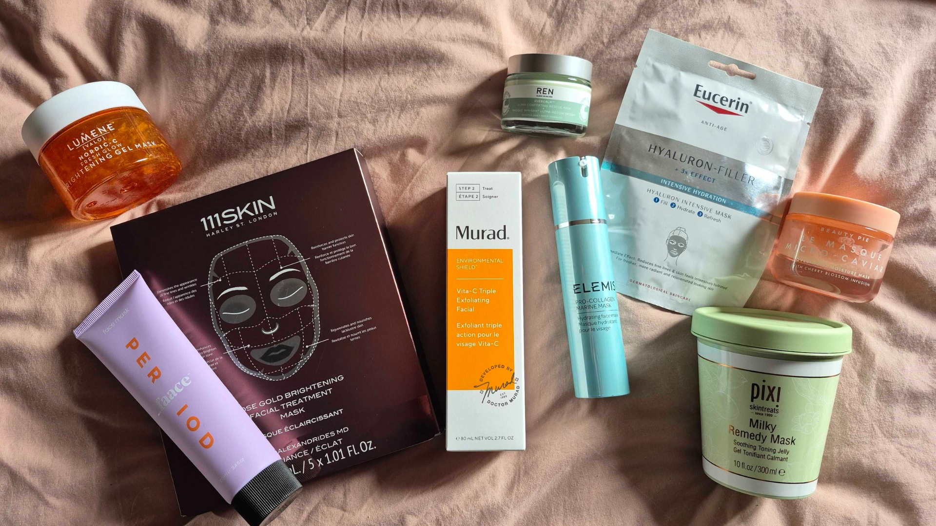 A selection of the best face masks featured in this guide, from brands including Lumene, 111SKIN, _Faace, Murad, REN, Elemis, Eucerin, Beauty Pie and Pixi, spread across a pink duvet cover