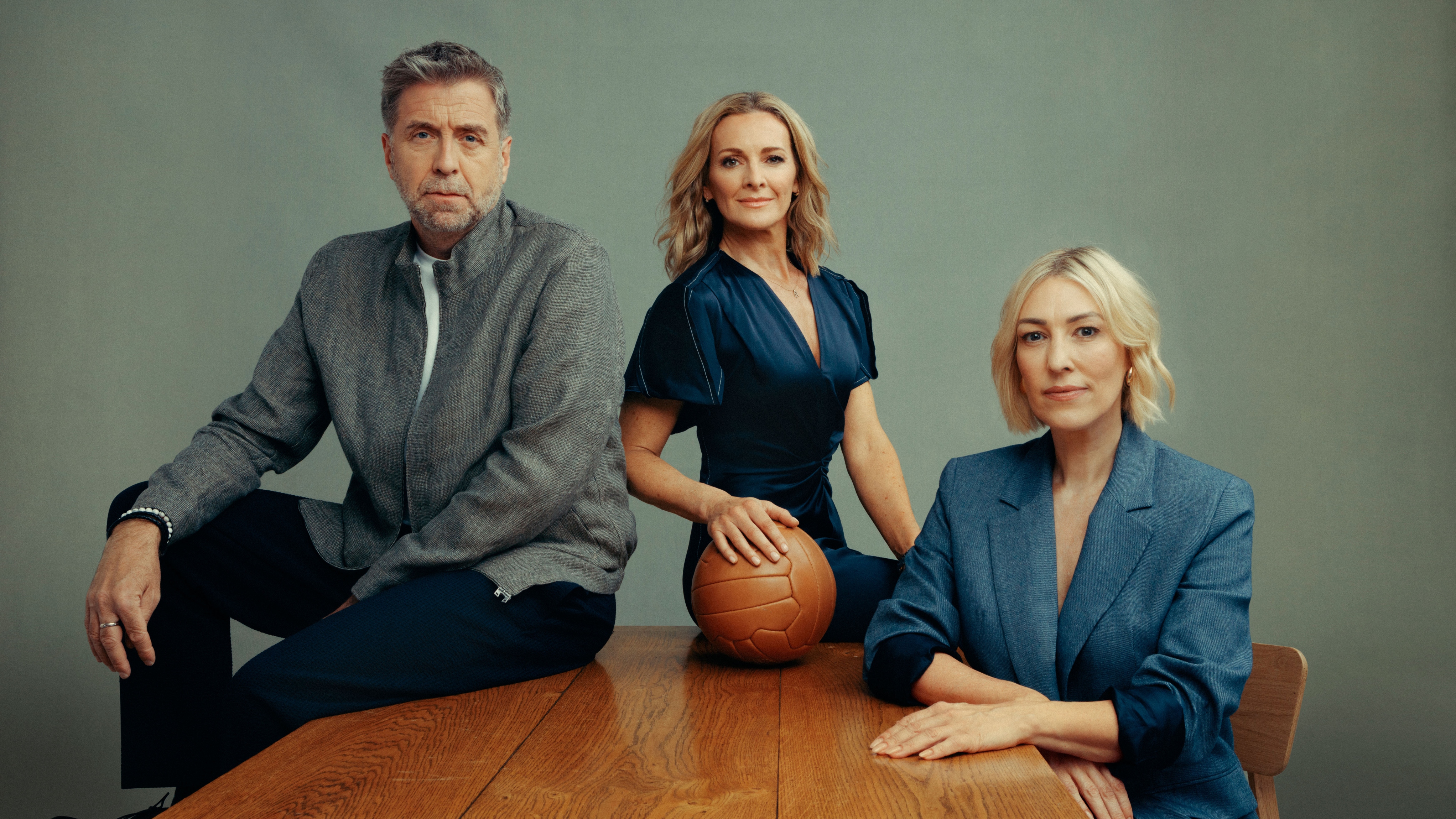 Gabby Logan, Mark Chapman and Kelly Cates sitting on a table in smart outfits - and Gabby is holding an old fashioned leather football