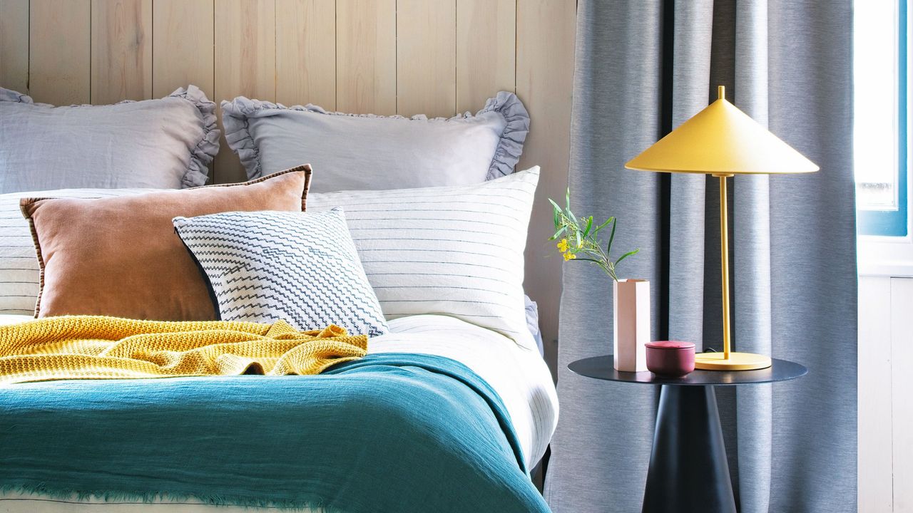 Bedroom with colourful bedding, bedside table, table lamp, and grey curtains