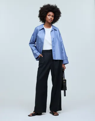 model wears blue cropped trench and black pants with a white t-shirt and black sandals while holding a black handbag