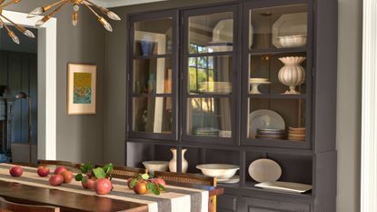 Dining room with a large dresser painted in black bean soup by Benjamin Moore, a wooden dining table with a striped runner and pomegranates on top, and grey walls. 