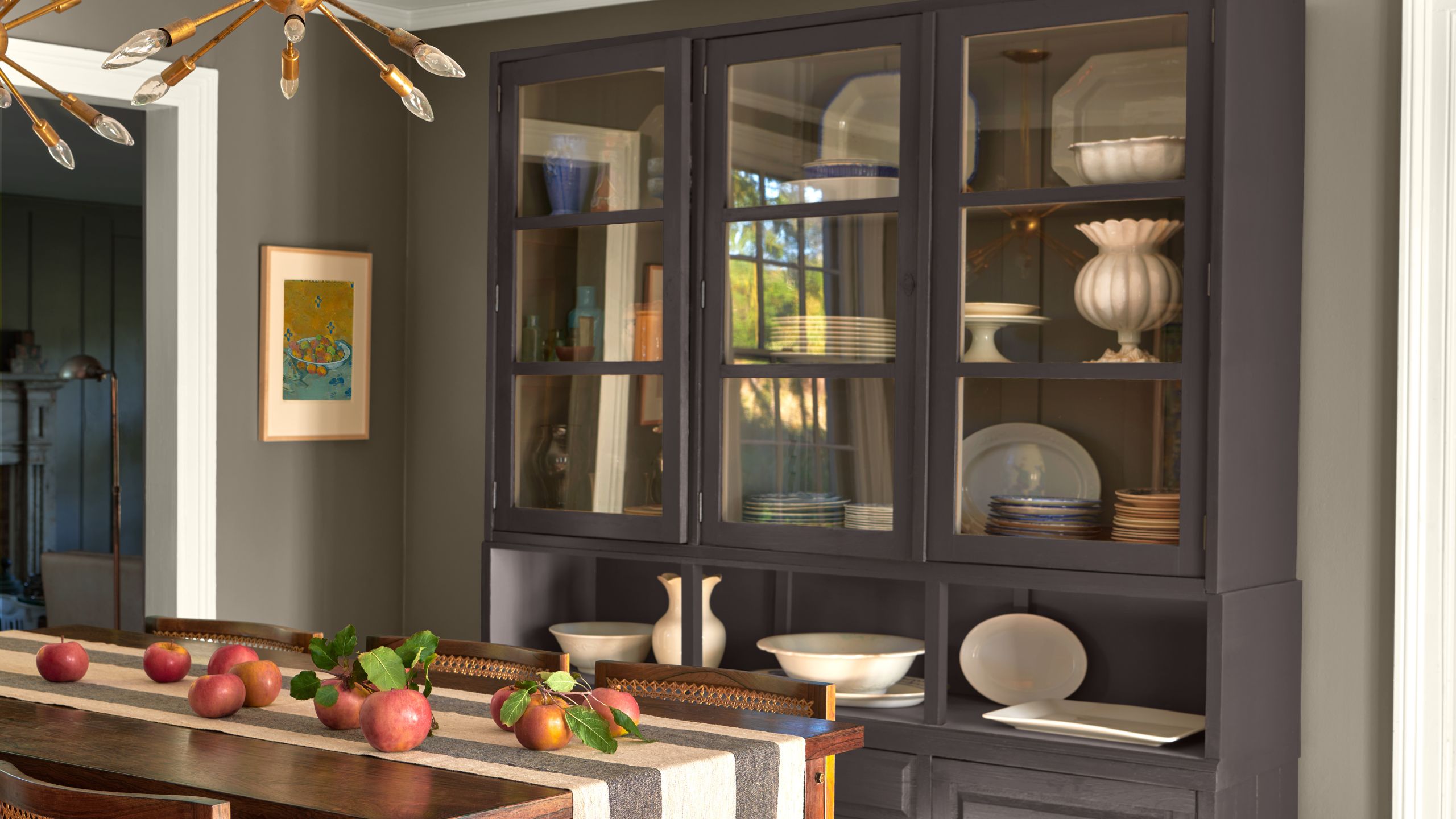 Dining room with a large dresser painted in black bean soup by Benjamin Moore, a wooden dining table with a striped runner and pomegranates on top, and grey walls. 