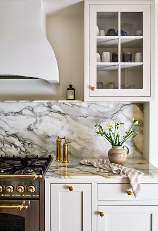 a white kitchen with glass cabinets next to a range cooker and hood. the cabinets are filled with mugs and glasses