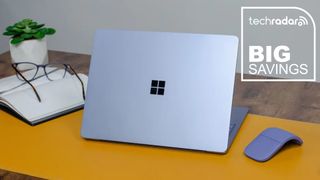 The Microsoft Surface Laptop in a home office during our tests next to a mouse, book, glasses, and plant, with a white TechRadar badge that reads 'Big Savings'