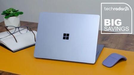 The Microsoft Surface Laptop in a home office during our tests next to a mouse, book, glasses, and plant, with a white TechRadar badge that reads 'Big Savings' 