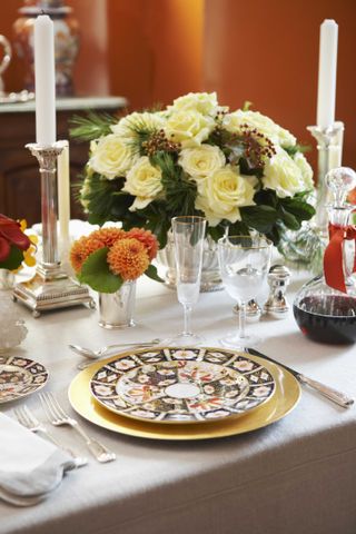 fall tablescape with cream roses and berries in vase, orange dahlia, gold rim glassware, patterned plates, silver candlesticks