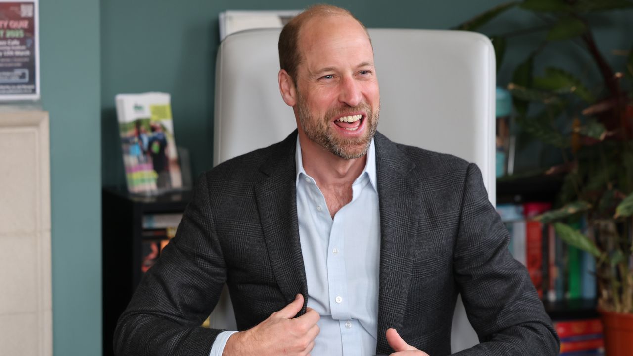 Prince William wearing a gray suit coat sitting in a chair and laughing 