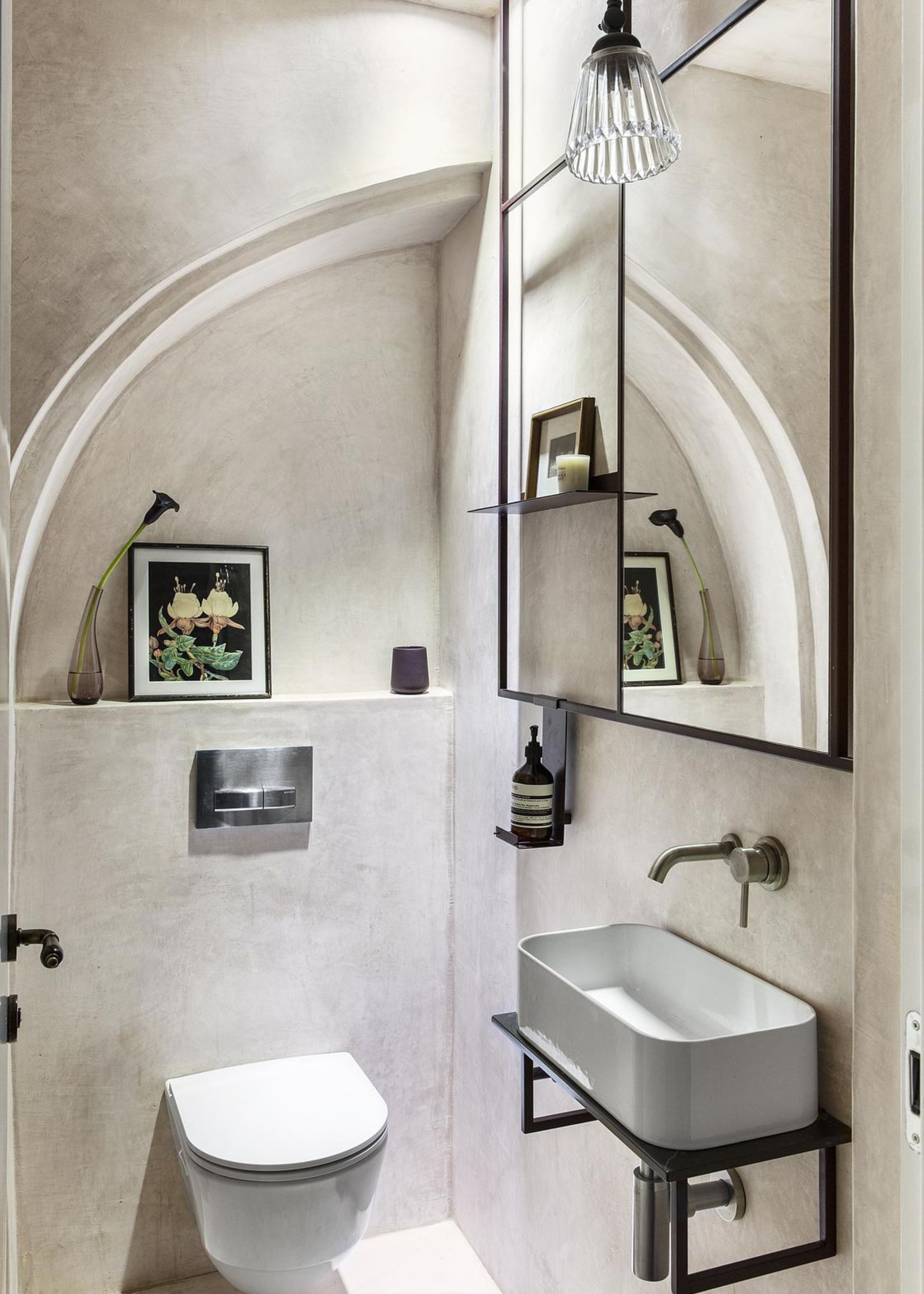 A small bathroom with micro cemented walls as well a view of the sink, mirror above the sink and a small toilet next to it.