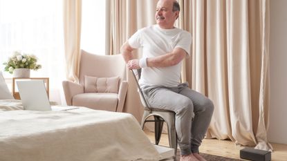 man in a white tshirt and grey joggers sitting in a chair and twisting to one side. he's in a bedroom setting with a laptop on the bed and a yoga block on the floor. 