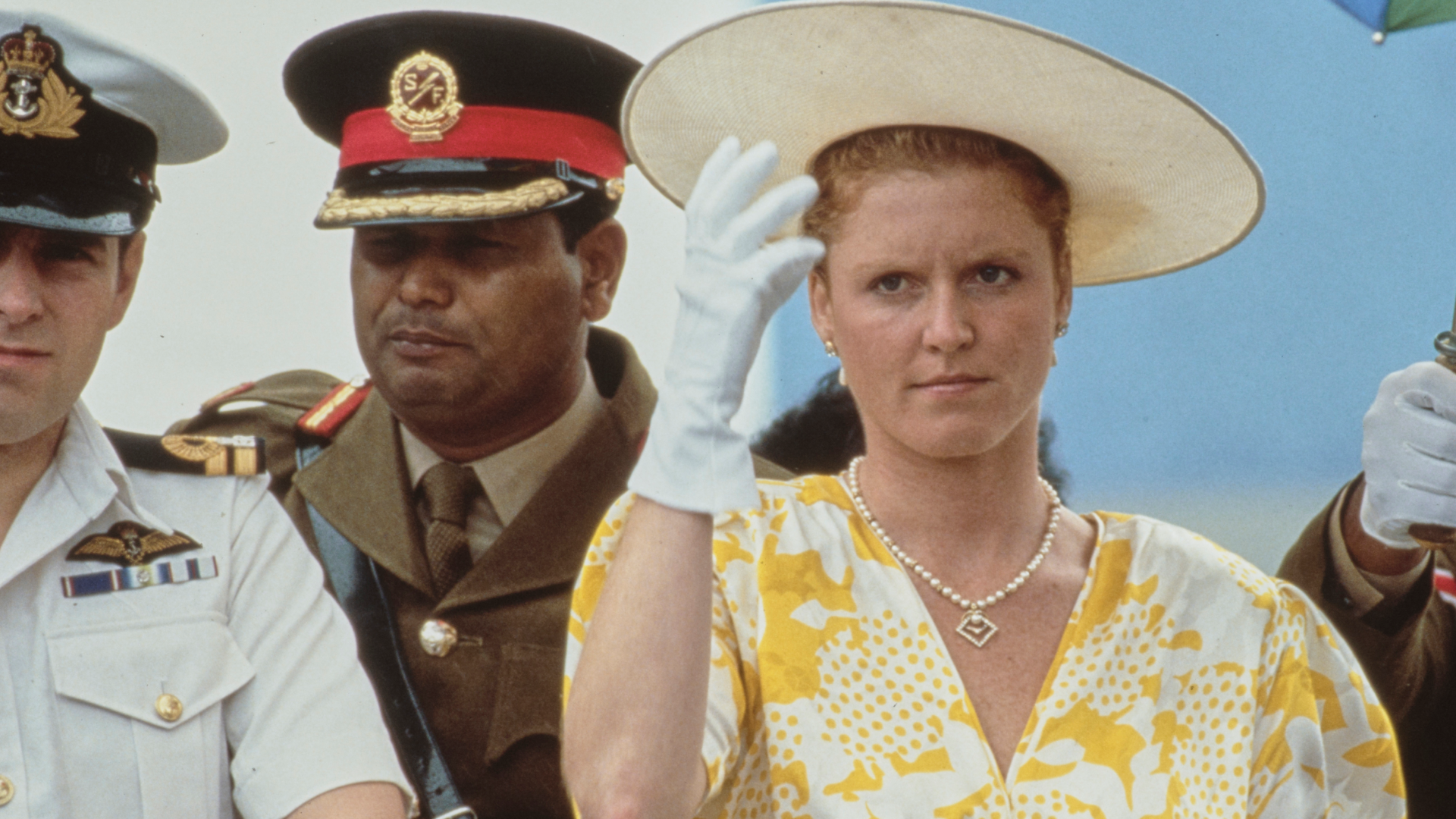 Sarah Ferguson scowling holding on to her yellow hat wearing a pair of white gloves, standing next to Prince Andrew, partially visible