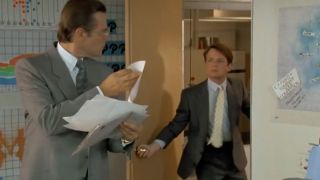 Michael J. Fox wearing a suit and walking into an office, surprising another man.