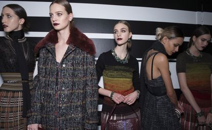 Five female models wearing looks from Bottega Veneta's collection. One model is wearing a black neck scarf and a black and multicoloured striped and plaid style piece. Next to her is a model wearing a grey coat with stripes in two different colours and maroon fur. Two models are wearing necklaces, black tops with a colour gradient design and red and white check style skirts. And the fifth model is wearing a grey and white sleeveless plaid style dress and dark coloured scarf